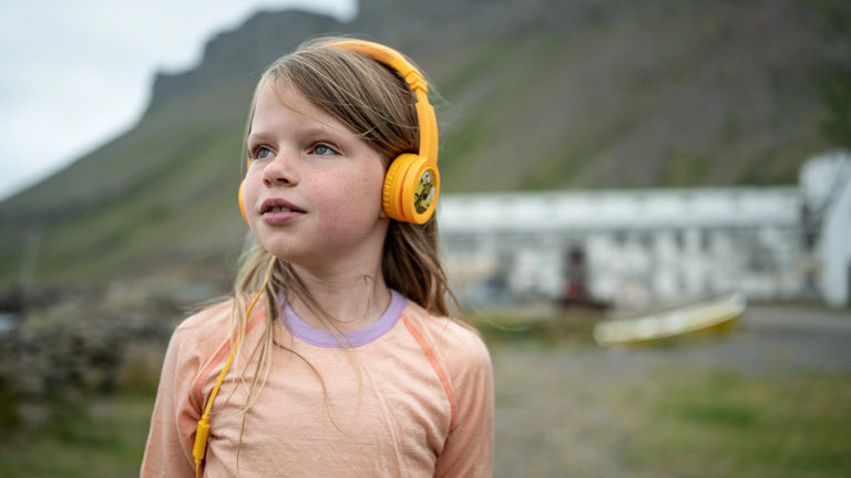 A young girl wearing yellow Onanoff BuddyPhones (headphones) and a light peach shirt stands outdoors with a blurred mountainous background.