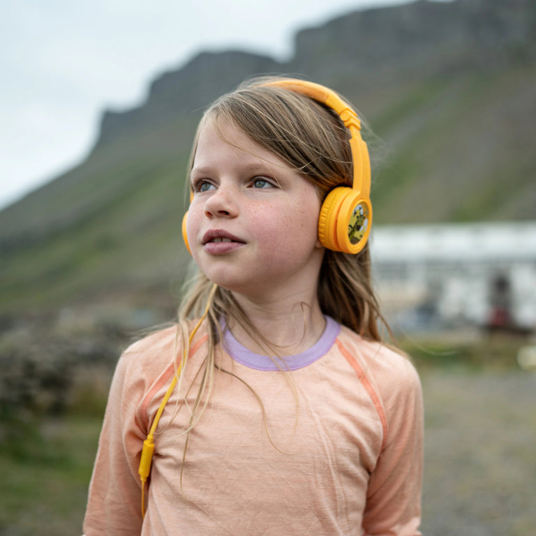 A young girl wearing yellow Onanoff BuddyPhones (headphones) and a light peach shirt stands outdoors with a blurred mountainous background.