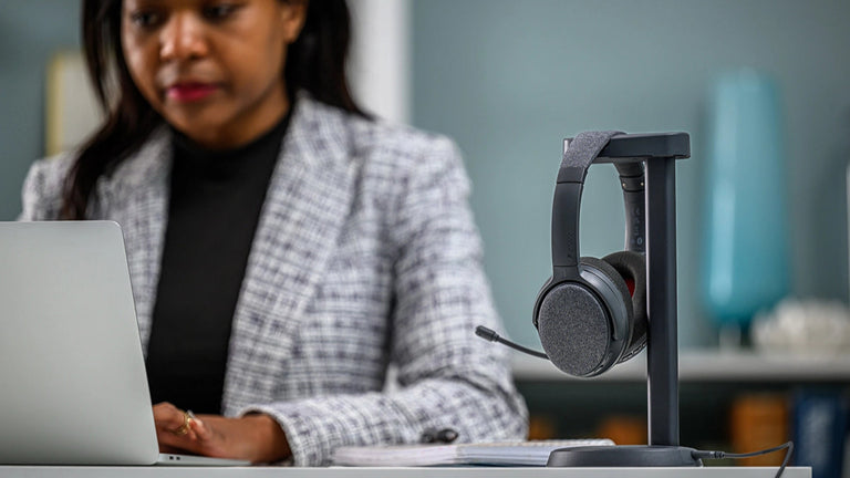 A woman in a checkered blazer sits at a desk with a laptop, beside a wireless charging docking stand holding a pair of Onanoff Fokus+ noise cancellation headphones.