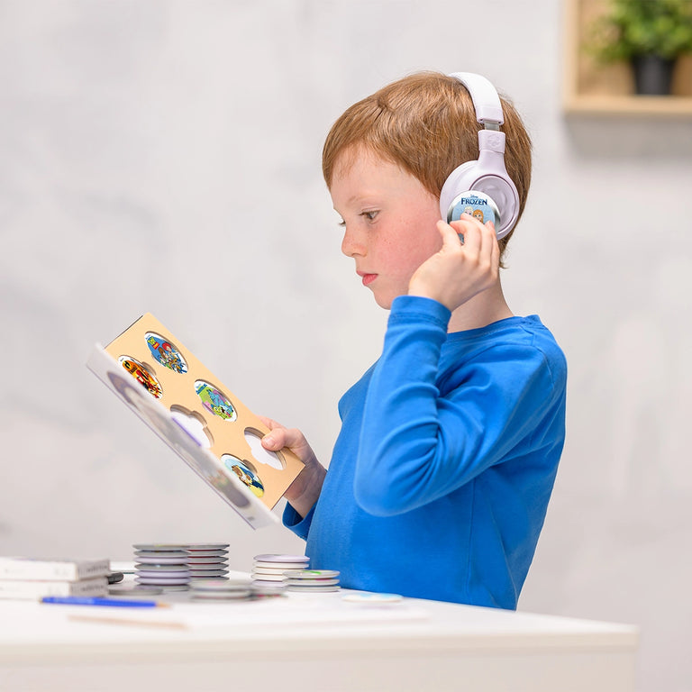 A young boy in a blue shirt wears headphones, possibly listening to Onanoff StoryPhones in gray, and looks at a StoryBook with StoryShields, standing at a white table indoors—immersed in playful learning.
