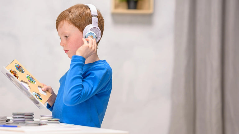 A young boy in a blue shirt wears headphones, possibly listening to Onanoff StoryPhones in gray, and looks at a StoryBook with StoryShields, standing at a white table indoors—immersed in playful learning.