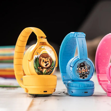 Colorful children's BuddyPhones (headphones) in yellow, blue, and pink, featuring fun cartoon stickers and soft cushions, displayed with books.