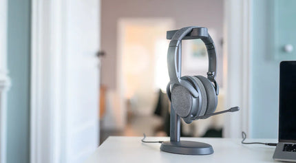 A gray Onanoff Fokus+ ANC headphones with a microphone rests on a sleek stand on a white desk, with a soft-focus background of a stylish room.