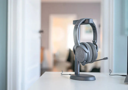 A gray Onanoff Fokus+ ANC headphones with a microphone rests on a sleek stand on a white desk, with a soft-focus background of a stylish room.
