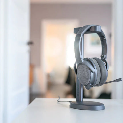 A gray Onanoff Fokus+ ANC headphones with a microphone rests on a sleek stand on a white desk, with a soft-focus background of a stylish room.