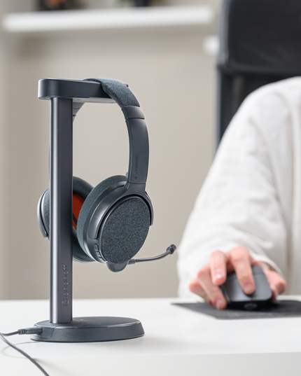 A pair of dusk gray ONANOFF Fokus+ wireless Bluetooth headphones on a wireless charging stand next to a computer.